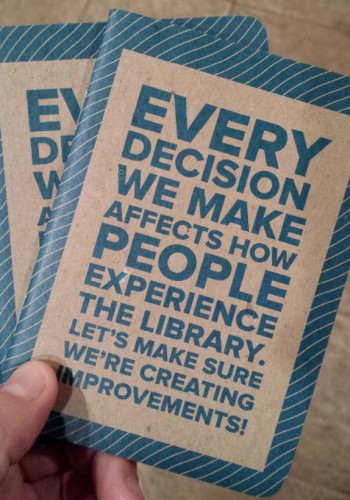 A hand holding 2 notebooks that say on the cover - every decision we make affects how people experience the library. let us make sure we're creating improvements.