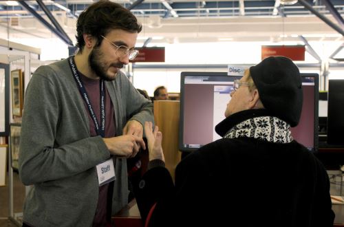 a library staff person answers a questions for a woman at a computer in the library.