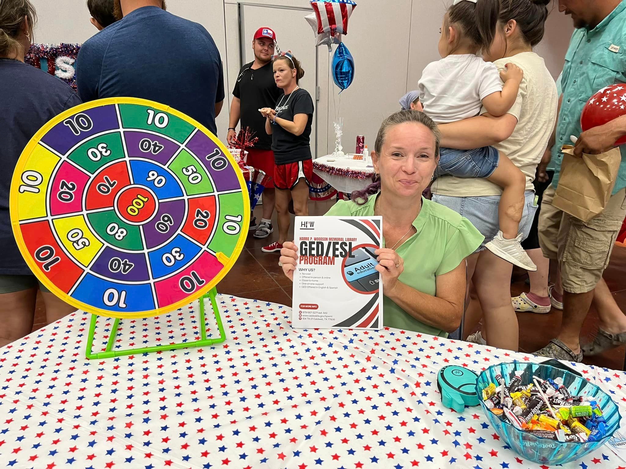 Volunteer sitting an information booth with festive decorations, holding a sign about GED and ESL programs