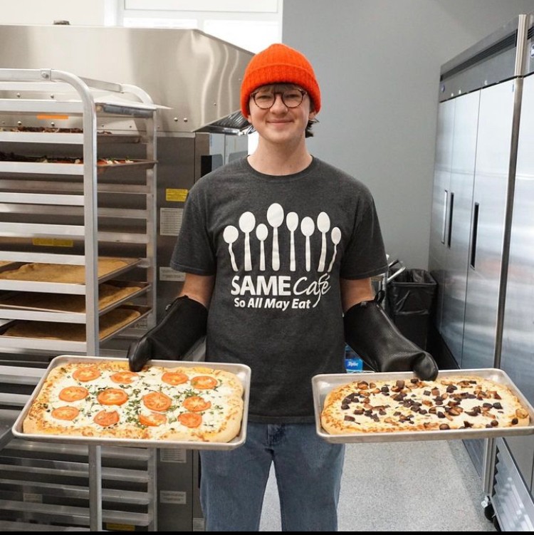 Person standing in a kitchen and holding two trays of pizza