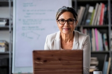 A woman sits at her laptop, smiling, with a whiteboard and bookshelves behind her.