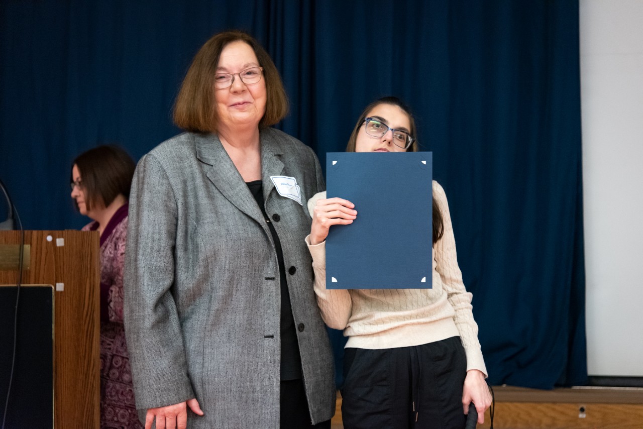 Two women standing in front of a stage, one of them holding an award
