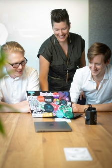 One standing adult between two seated adults looking at a laptop