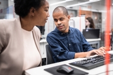 Two colleagues discussing work at a desk in a modern office environment, with one person typing on a keyboard and a computer mouse visible on the desk.