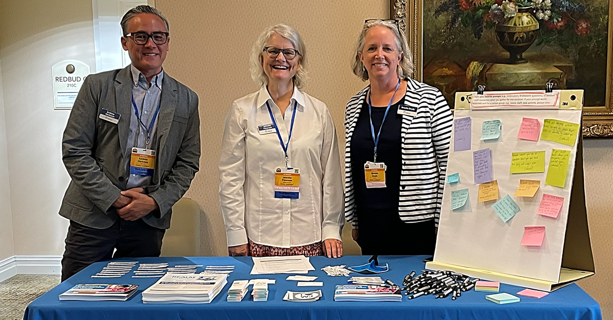 WebJunction team members (l to r) Andrew Harbison, Jennifer Peterson, and Brooke Doyle standing in front of a booth decorated with a WebJunction tablecloth